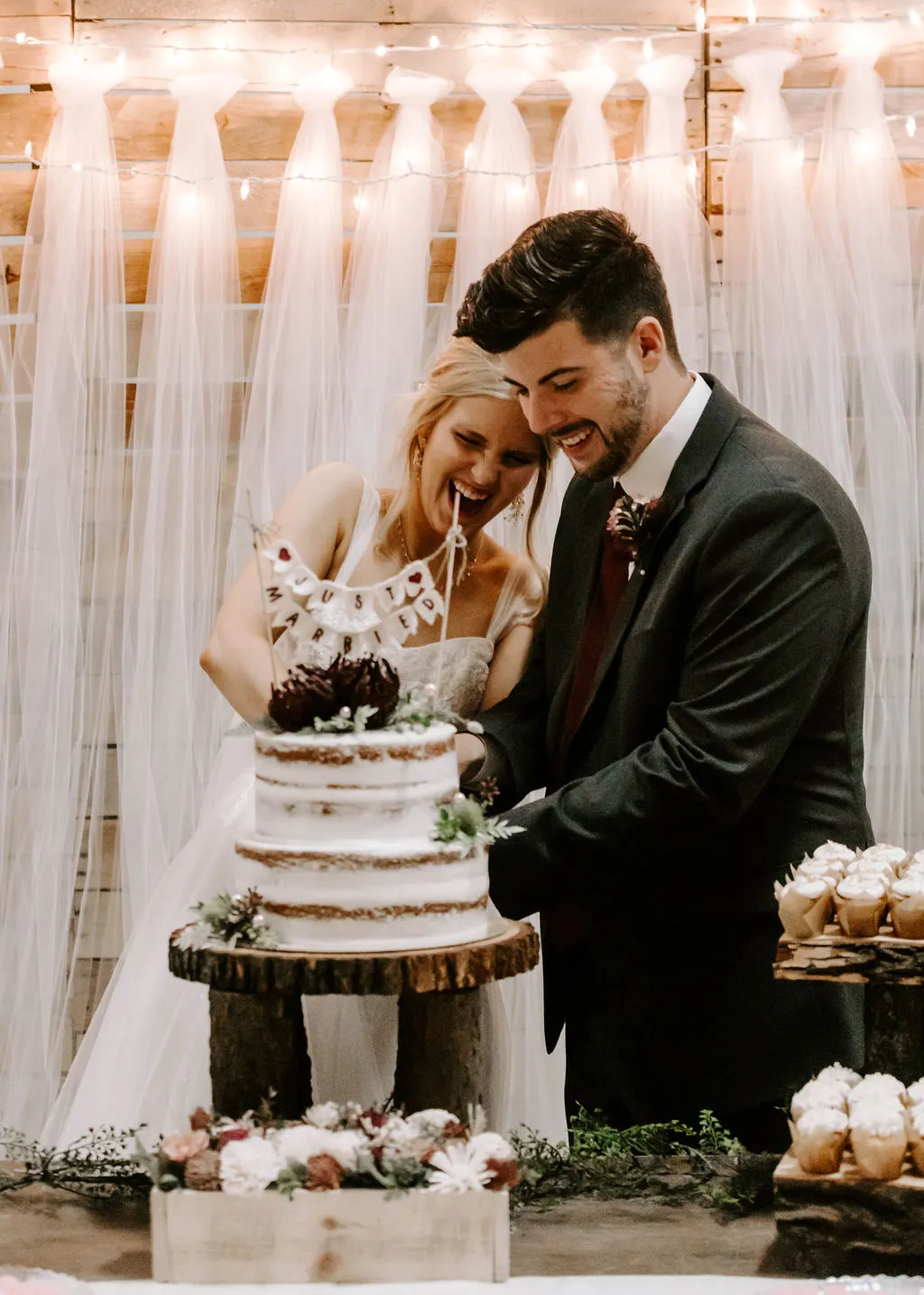 Bride and groom cutting their wedding cake in front of a draped tulle and string-light backdrop